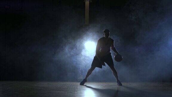 一名男子在黑暗的篮球场上拿着篮球背景是烟雾中的篮球圈在三盏背光灯的照耀下他展示着运球技巧