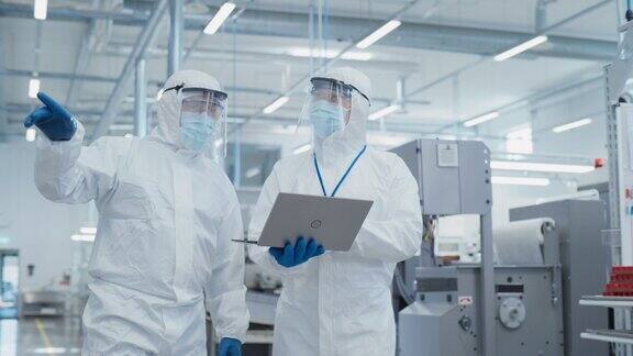 两名科学家穿着无菌工作服和口罩站在重工业工厂使用笔记本电脑检查工业机器设置和配置生产功能