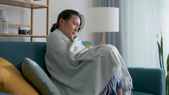 亚洲女人觉得冷裹着毯子坐在家里客厅的沙发上