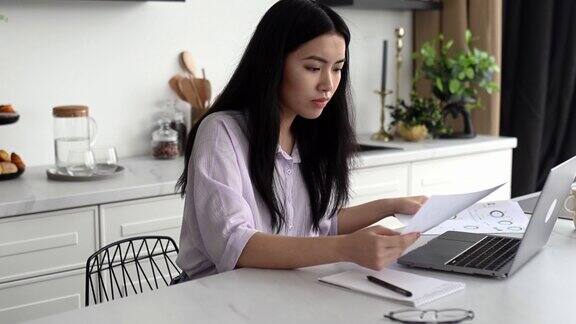 集中的聪明的深色头发的中国女孩穿着时髦的衣服自由职业者学生或创意设计师坐在家里的厨房里使用笔记本电脑在一个项目上工作浏览互联网分析文件