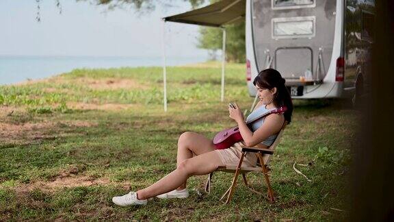 年轻的亚洲女子使用智能手机和她的尤克里里在露营车在海滩露营车旅行系列