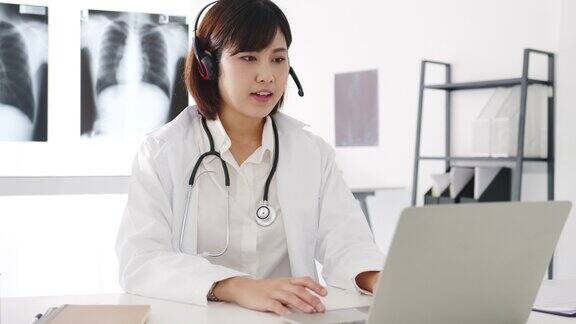 年轻的亚洲女医生身穿白色医疗制服带听诊器使用电脑笔记本电脑在诊所或医院的办公桌上与病人进行视频电话会议