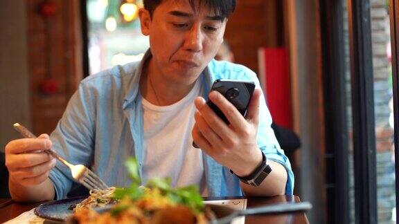 年轻的亚洲男子一边吃面条一边看手机
