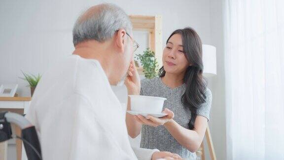 亚洲迷人的年轻女儿在家里为年长的父亲提供食物美丽的孙女支持和照顾老人坐在轮椅上的老人在客厅医疗保险