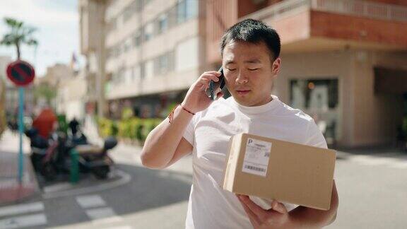 年轻的中国快递员在街上拿着包裹在智能手机上说话