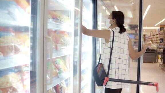 年轻的亚洲女性在超市的冷冻室里选择和挑选产品戴上防护灰尘和病毒的面罩保持健康