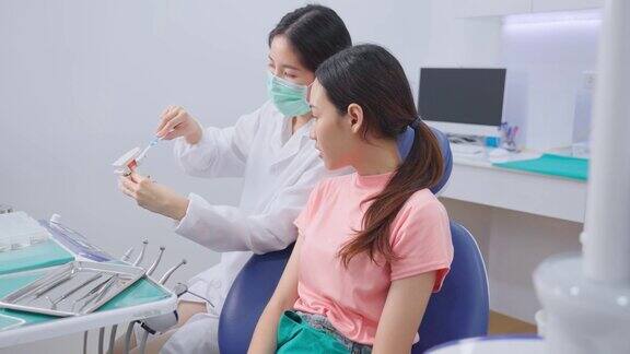 亚洲年轻女孩正在听女牙医讲解口腔护理和牙齿矫正患者的刷牙牙医在牙科诊所佩戴防护口罩预防COVID-19