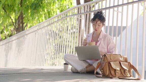 女商人一边吃面条一边用笔记本电脑在公园里工作