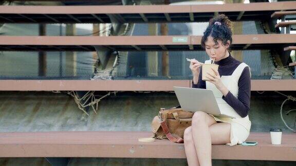 一位女商人一边吃面条盒饭一边用笔记本电脑在公园里工作
