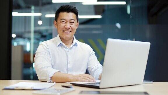 快乐的亚洲男性员工商人或会计律师公证人银行家在笔记本电脑上工作微笑看着相机坐在办公室的办公桌前