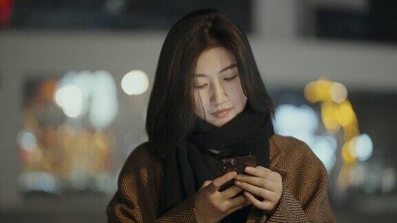 一个年轻漂亮的亚洲女人在夜晚用手机背景是模糊的霓虹灯4k