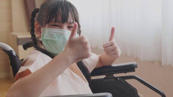 在新冠肺炎大流行期间坐在轮椅上戴口罩的亚洲小病童孩子感觉良好充满希望高兴地竖起大拇指