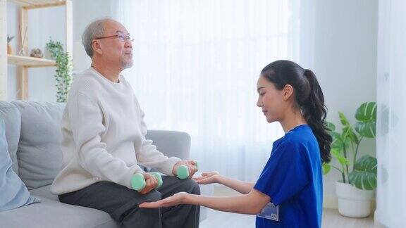 亚洲年长男子在护士的支持下做物理治疗老年男性患者坐在客厅的沙发上使用哑铃锻炼与治疗师医生在养老院照顾