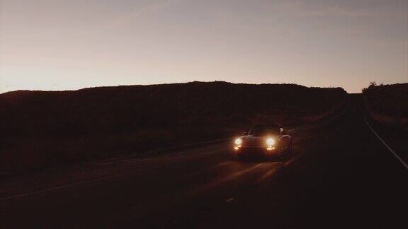 鸟瞰图红色经典汽车沿着荒凉的沙漠道路在黄昏与头灯打开