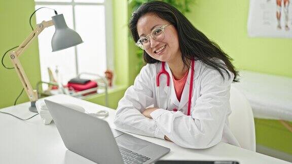 年轻的中国女医生双手交叉坐在诊所的桌子上使用笔记本电脑