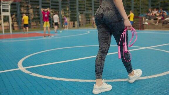 运动女孩在篮球场上走着她拿着一根跳绳和一瓶水背景是人们在打地板球4k