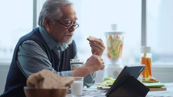 亚洲男性老年白发吃健康早餐面包和注入水阅读新闻社会问题的平板设备
