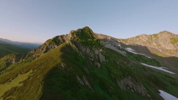 FPV运动无人机飞越覆盖着绿色草地和融化的雪的岩石山脉