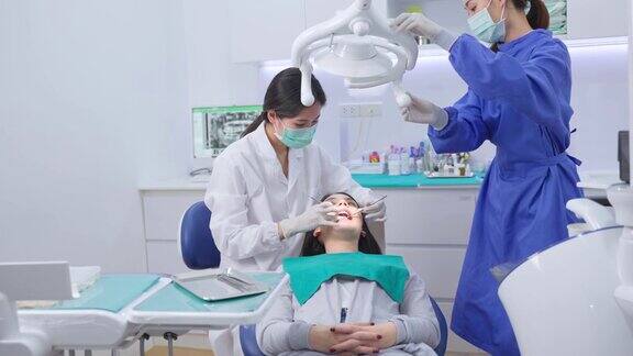 亚洲女牙医调整牙科手术灯然后开始检查或检查年轻女孩病人的牙齿躺在牙科椅上牙科助理在牙科诊所提供器械支援