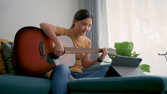 一个亚洲女人拿着吉他坐在沙发上在家里的客厅里用平板电脑学习吉他