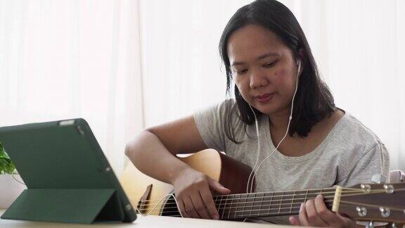 亚洲女子白天坐在靠窗的家里用平板电脑在线练习弹原声吉他具有技能提升理念的周末活动