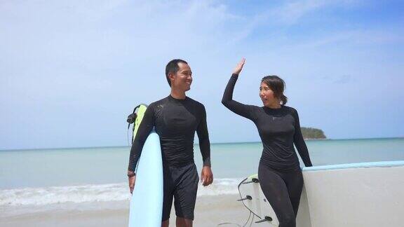 一对亚洲夫妇穿着潜水服手拿冲浪板一起在海滩上散步