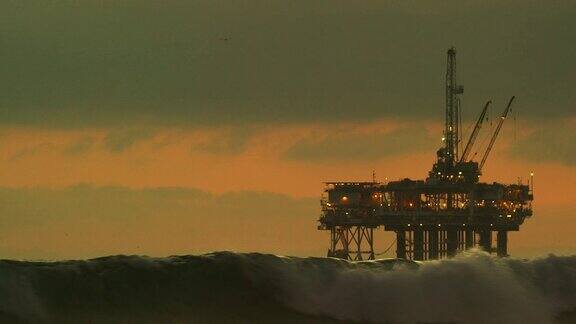 在一个戏剧性的暴风雨的天空下的日落下海浪撞击在地平线上的近海石油钻井平台在南加州亨廷顿海滩海岸