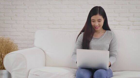 年轻的亚洲女子坐在家里客厅的沙发上使用笔记本电脑