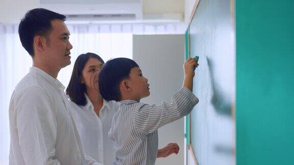 亚洲可爱的儿子在教室里和家长一起在黑板上画画小男孩在幼儿园做有趣的教育活动在黑板上写字母和数字和爸爸妈妈一起玩