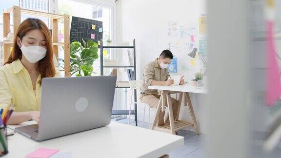 新常态下亚洲女性在办公室使用笔记本电脑和丙烯酸隔板站隔开时戴着口罩进行社交距离以防病毒