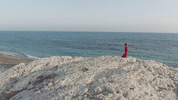 在塞浦路斯地中海的悬崖上有才华的女人拉小提琴自信的白人音乐家身穿红裙在日落时分欣赏户外音乐自然与艺术观念
