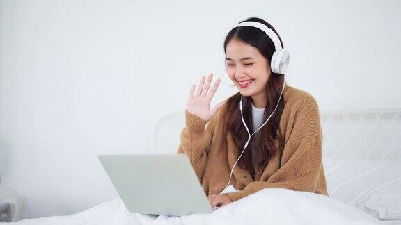快乐美丽的年轻亚洲女人工作在电脑笔记本电脑和坐在床上在房子她一边开视频会议一边戴着耳机听音乐