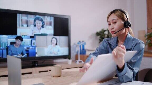 年轻的亚洲女商人戴着耳机使用笔记本电脑进行视频会议电话向同事展示财务计划同时在家庭办公室的会议室工作