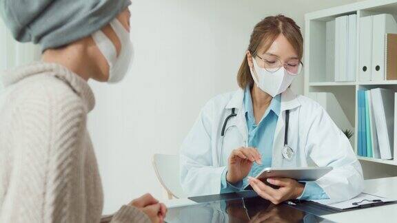 年轻的亚洲女医生戴着防护口罩在卫生所用平板电脑为患癌症的女病人看病