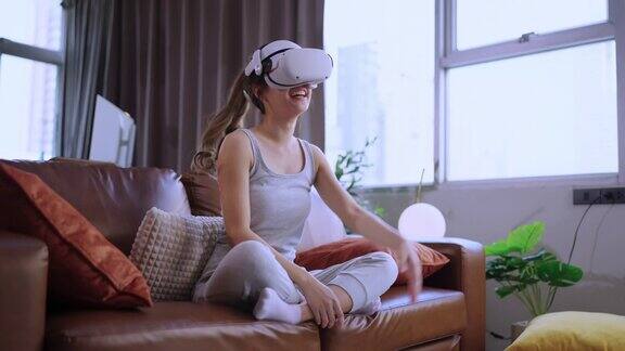 年轻迷人的休闲亚洲女性通过VR眼镜头戴护目镜在沙发上观看了虚拟现实中的表演直播音乐会在家里的客厅里享受着乐趣和快乐的时刻