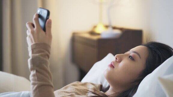 一名亚洲华裔妇女躺在床上盖着毯子患感冒和流感用智能手机和朋友聊天用数字体温计测量体温