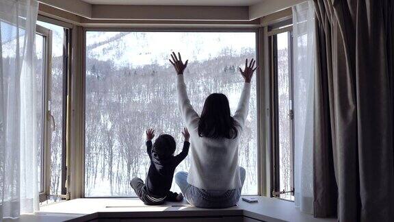 一个小孩和他的母亲坐在窗边看着森林里的雪花飘落
