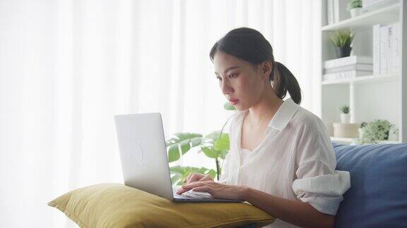 年轻的亚洲妇女工作的笔记本电脑而坐在家里的客厅沙发