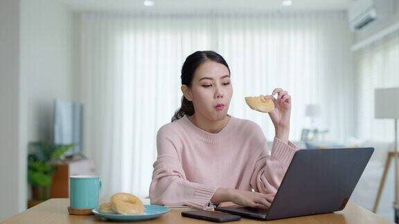 年轻迷人美丽的亚洲女性饿了吃甜甜圈吃零食吃满嘴看电脑笔记本在家里在忙碌的工作从家里多任务不健康的饮食生活理念