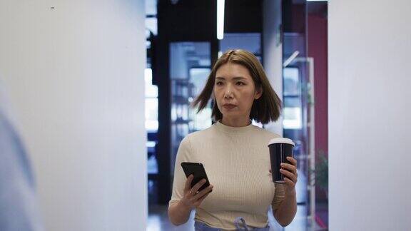 在现代办公室里拿着咖啡杯拿着智能手机走路的亚洲女商人