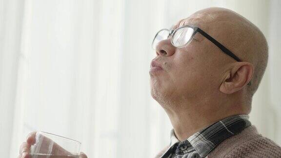 一位中国老人站在窗前一手拿着一杯水正在吃药