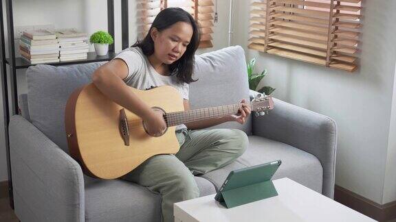 一名年轻的亚洲女子坐在沙发上用家里的笔记本电脑在线学习吉他演奏