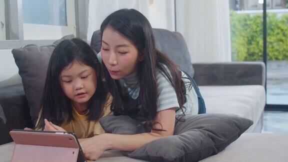 年轻的亚洲家庭和女儿喜欢在家里使用平板电脑日本妈妈和小女孩躺在家里客厅的沙发上看电影有趣的妈妈和可爱的孩子玩得很开心