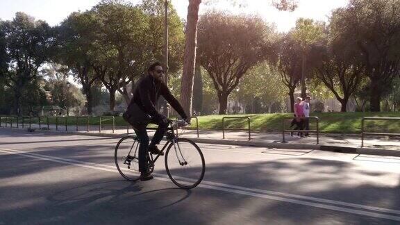 在阳光明媚的日子里年轻的潮男在罗马市中心一条有树的道路上骑着自行车戴着太阳镜