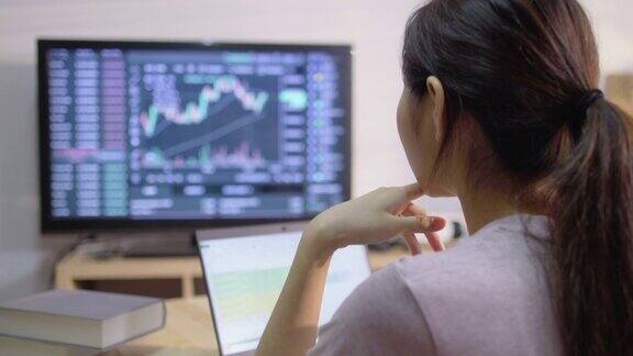 年轻的成年女性坐在家里的办公室一边用笔记本电脑工作一边在电脑屏幕上看股票市场数据