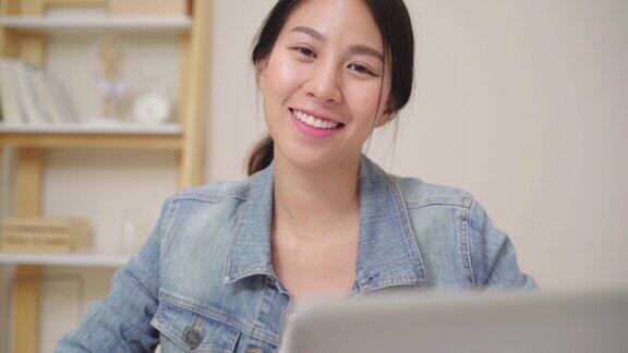 亚洲商业女人感到幸福的微笑和看着相机而放松在家里的办公室年轻的亚洲女性在家里客厅的桌子上使用笔记本电脑工作