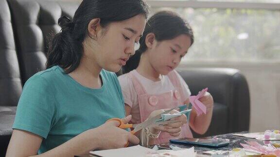 两个亚洲姐妹女孩在家的时候在客厅里剪纸和做工艺品姐姐用彩纸、剪刀和胶水在纸上创作艺术作品形成她的想象艺术和工艺概念