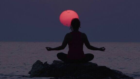 在令人惊叹的日落天空和大太阳期间坐在海水中的石头上冥想的女人的剪影治疗身体心理和精神状态的人