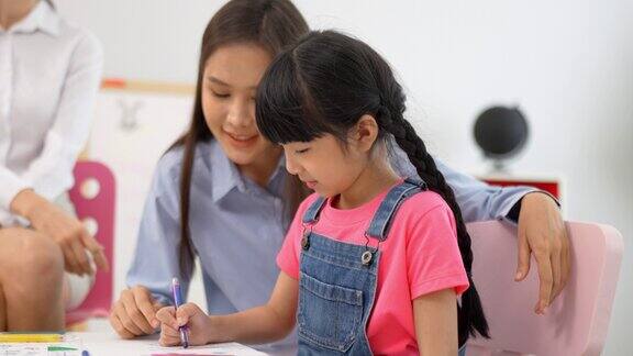 幼儿园教育学校女老师在教小女孩在教室的桌子上画彩绘本多民族幼儿教师和学生的课堂幼儿园和学习理念