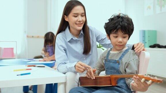年轻的亚洲老师在教他的小男孩吉他和尤克里里小男孩在幼儿园学吉他特写学校里的尤克里里琴课孩子跟老师学吉他音乐教育